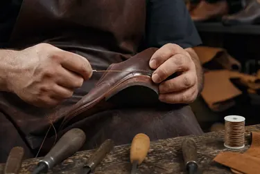Sapateiro costurando um calçado, mostrando detalhes da costura e ferramentas.